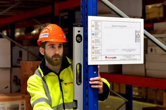 Pracownik w kasku i szelkach bezpieczeństwa wykonujący prace konserwacyjne na wysokim regale magazynowym z zabezpieczeniami